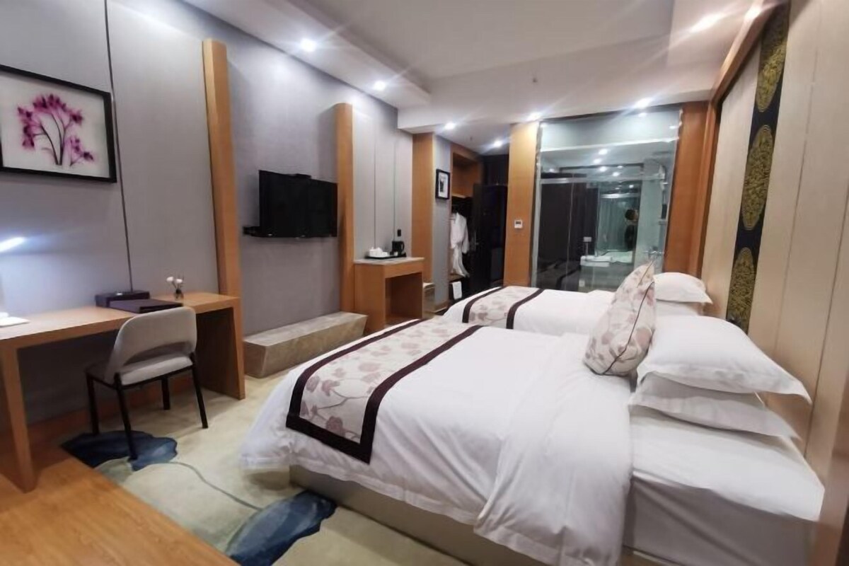 Ein Hotelzimmer mit zwei Betten, einem Schreibtisch, einem Stuhl, einem Fernseher und einem durch eine Glastür einsehbaren Badezimmer.