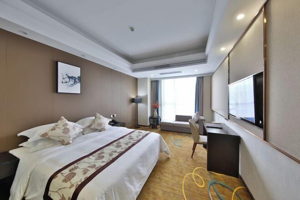 Ein Hotelzimmer mit einem großen Bett, einem Schreibtisch, einem Sessel, einem Fernseher und einem Fenster mit Vorhängen.