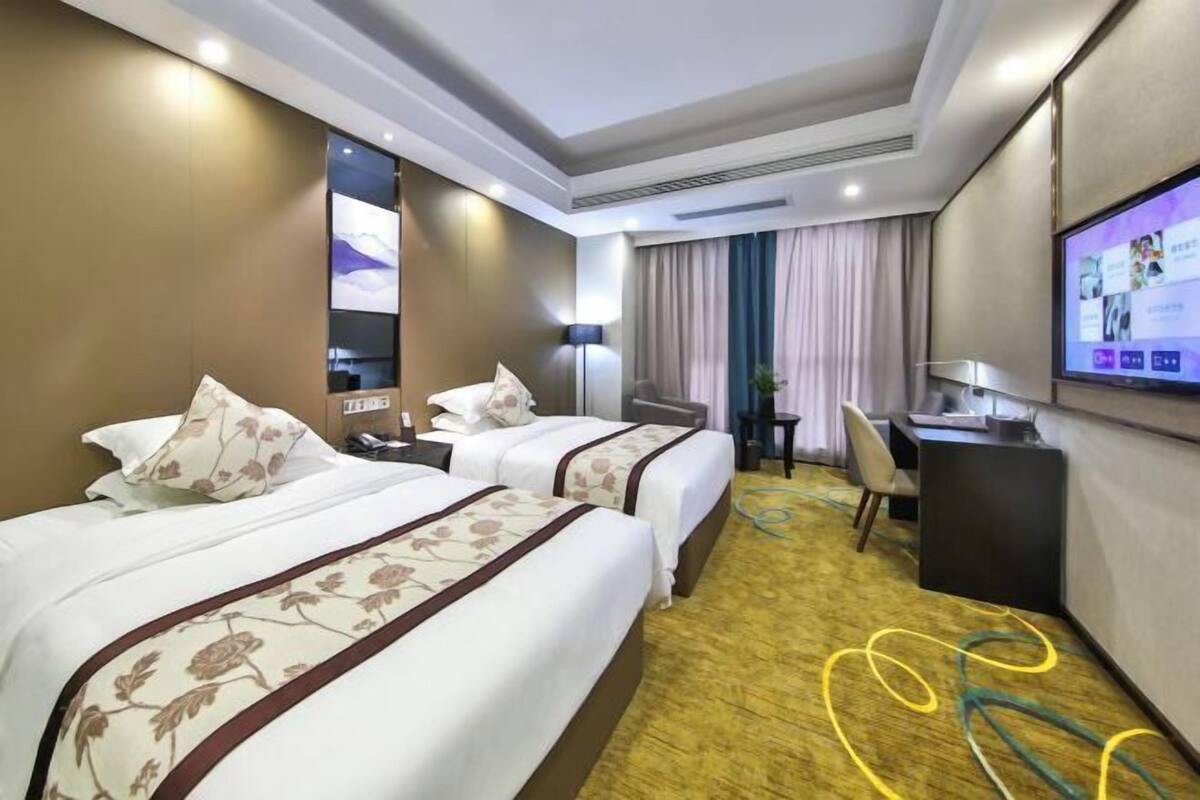 Ein Hotelzimmer mit zwei Betten, einem Schreibtisch, einem Stuhl, einem Fernseher und einem großen Fenster mit Vorhängen.