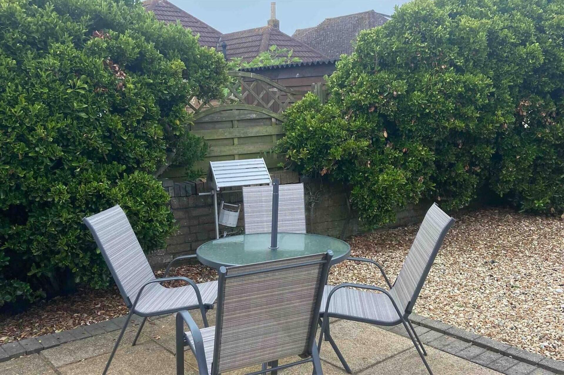 Een terras met een ronde tafel en vier stoelen, omringd door dichte groene struiken en een houten hek.