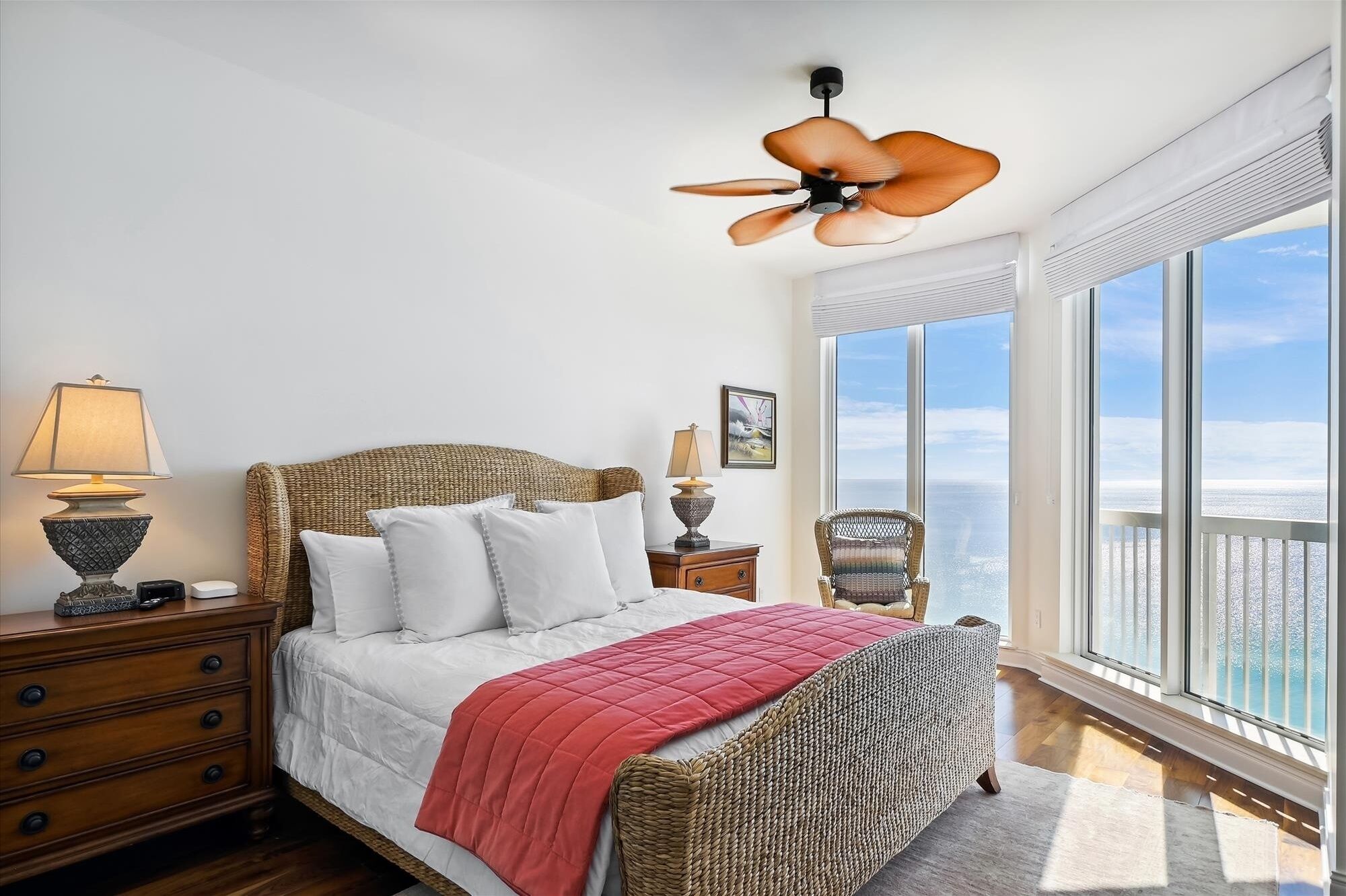 A bedroom with a bed, a nightstand, a lamp, a ceiling fan, and a large window with a view of the ocean.