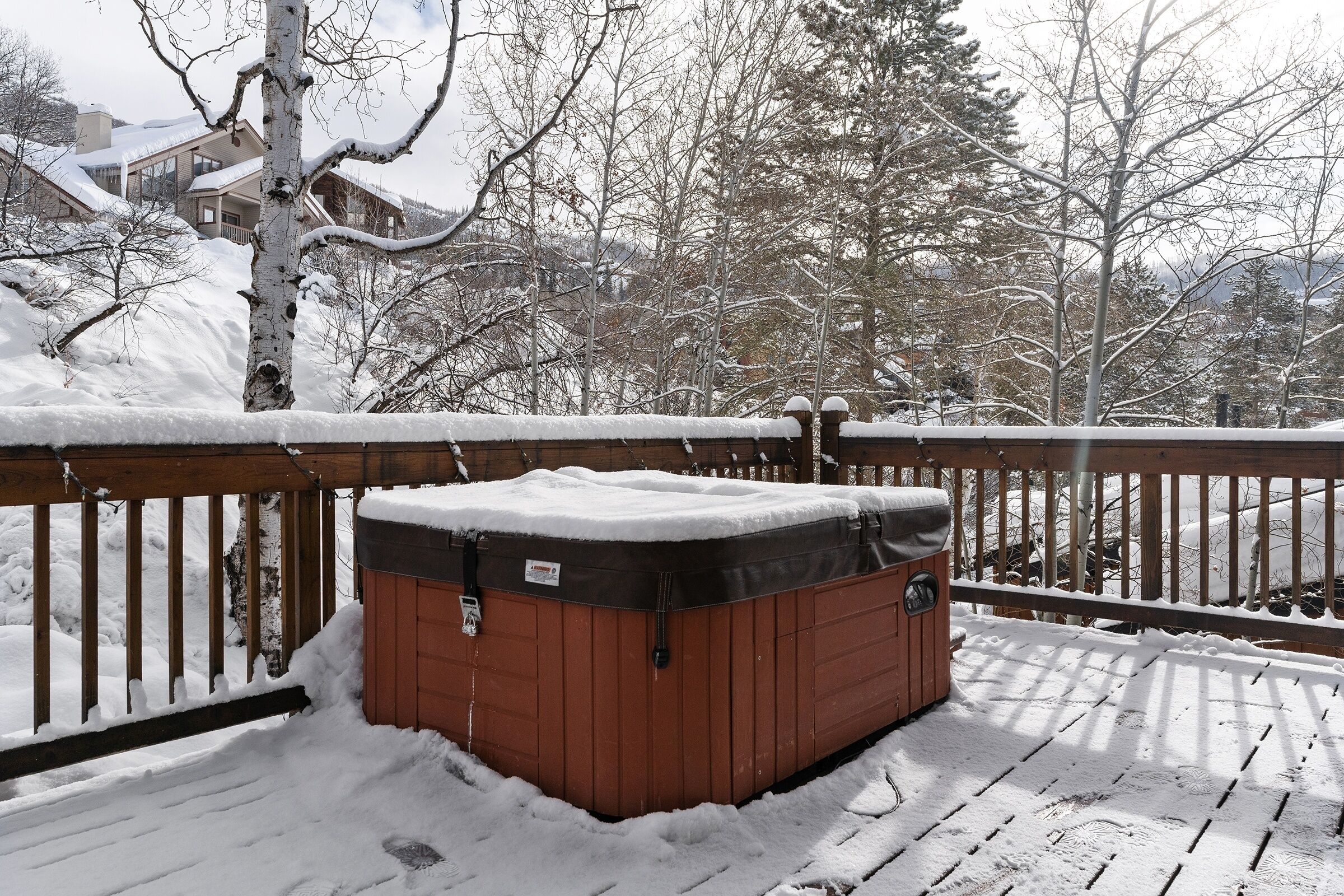 Un jacuzzi en una terraza cubierta de nieve, con barandilla de madera y árboles nevados al fondo.