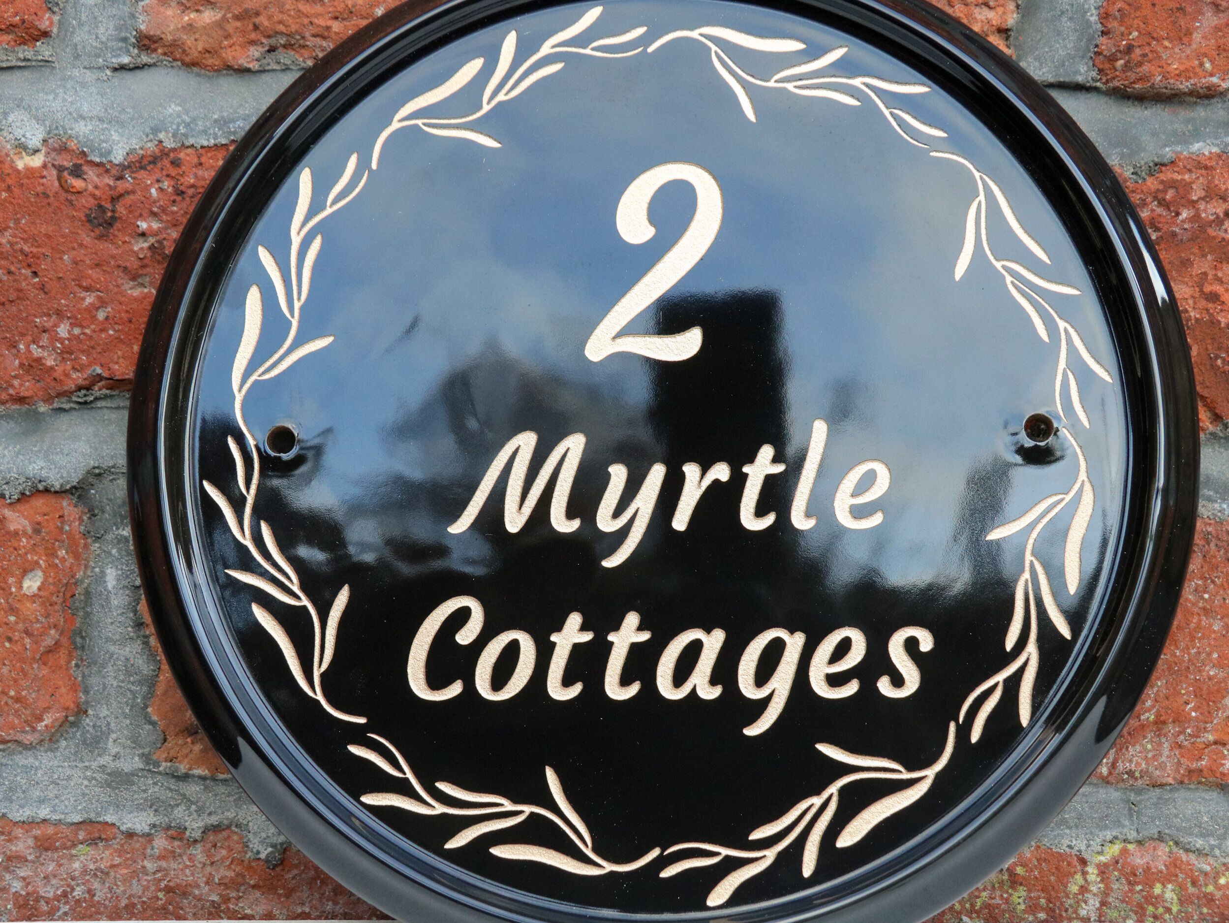 Un senyal rodó negre amb lletres i elements decoratius blancs que diu "2 Myrtle Cottages".