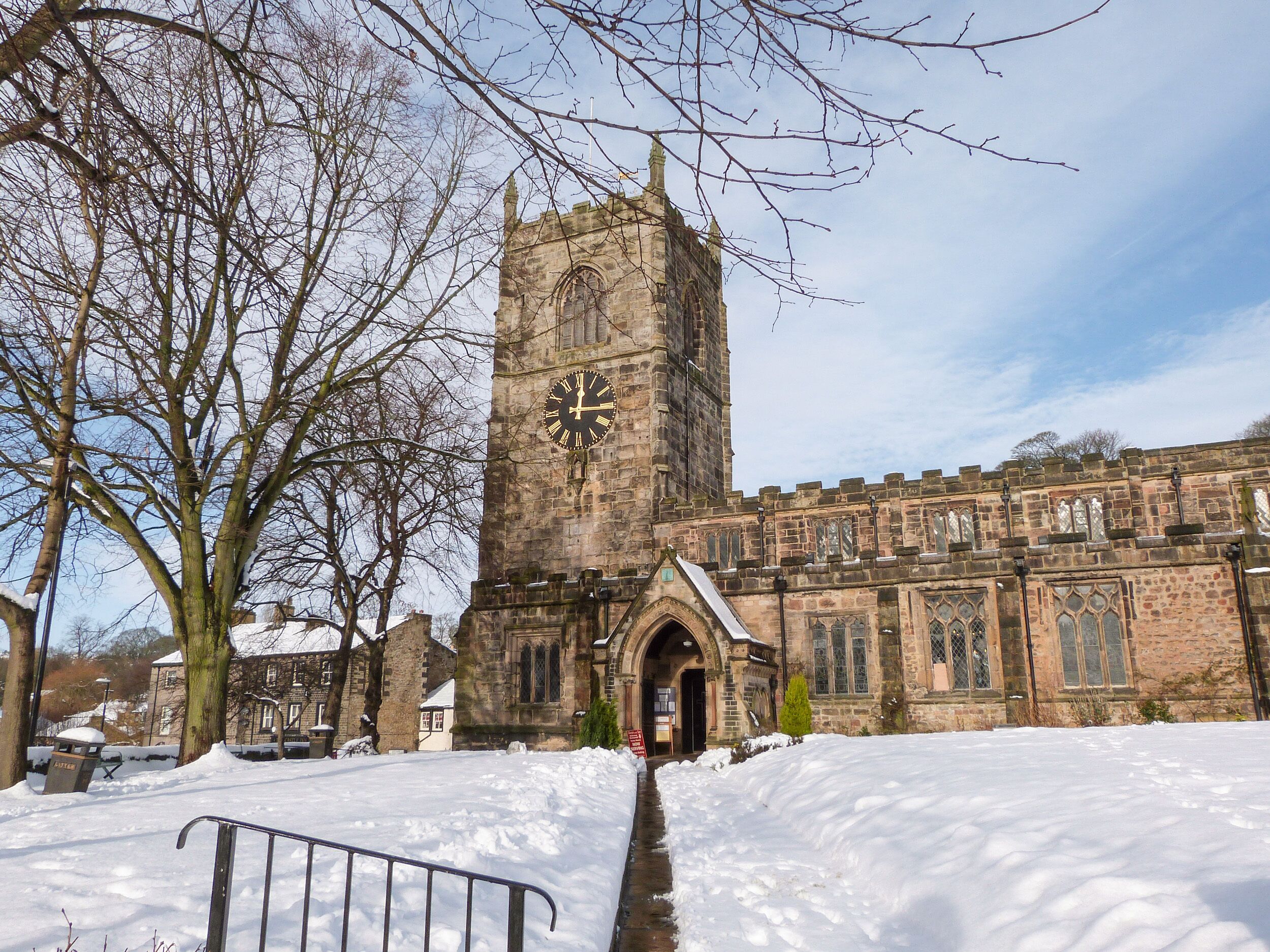 A historic stone building with a prominent clock tower, a snow-covered pathway, and a large tree in the foreground.