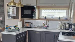A modern kitchen with dark grey cabinets, a black microwave, a stainless steel kettle, and a window with a view. - 8 Birth Static Caravan (Filey)