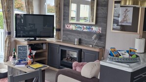 A modern living room with a flat-screen TV, a fireplace, a dining area with a table set for two, and a kitchen area with a sink and countertop. - 8 Birth Static Caravan (Filey)