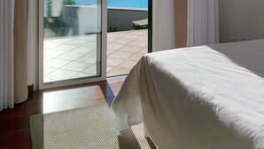 Ett sovrum med en säng, utsikt över havet genom en skjutdörr och en uteplats med kakelplattor.