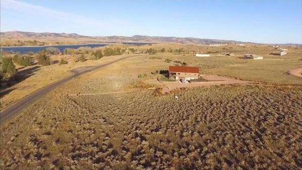 A rural landscape with a house surrounded by dry vegetation and a lake in the background.