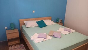 Un letto singolo con materasso verde, cuscini bianchi e color acquamarina, e asciugamani piegati in tonalità di viola e bianco. Accanto al letto, un comodino in legno con una lampada verde.