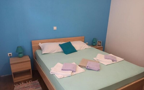 Ein Einzelbett mit einer grünen Matratze, Kissen in Weiß und Türkis sowie Handtücher in Lila- und Weißtönen. Neben dem Bett steht ein Nachttisch aus Holz mit einer grünen Lampe.