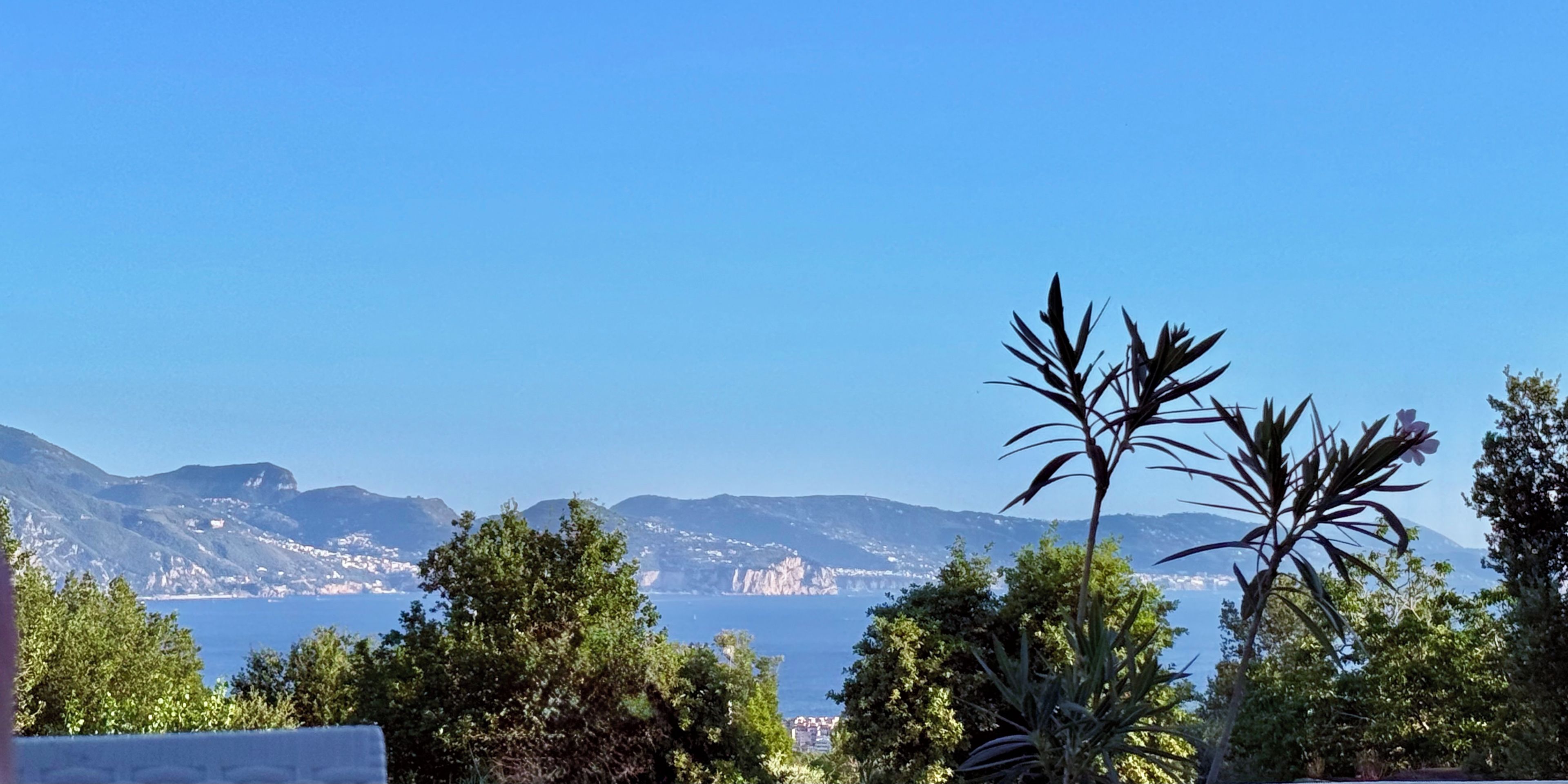 A coastal landscape with mountains, a clear sky, and dense greenery.