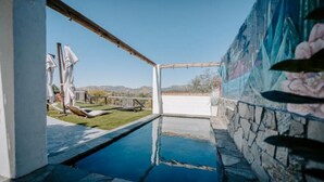A swimming pool with a stone wall and a mural, surrounded by a patio area with lounge chairs and umbrellas. - Vibey😎Pool House🏡 ModernFarmhouse 1700sqft + View🌄 (Lakeside)