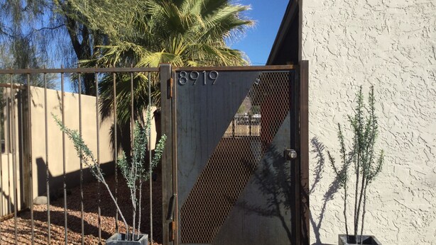 A gated entrance with a metal gate and a number plate, surrounded by potted plants and a palm tree. - Urban escape,Next to shopping center and restaurants! (Phoenix)