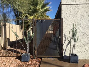 A gated entrance with a metal gate and a number plate, surrounded by potted plants and a palm tree.