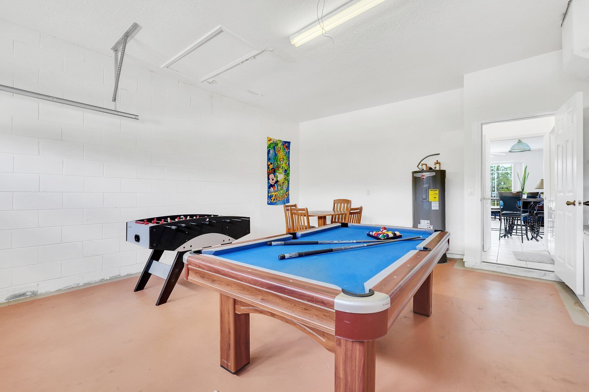 A pool table with a blue felt surface, a foosball table, and a colorful poster on the wall.