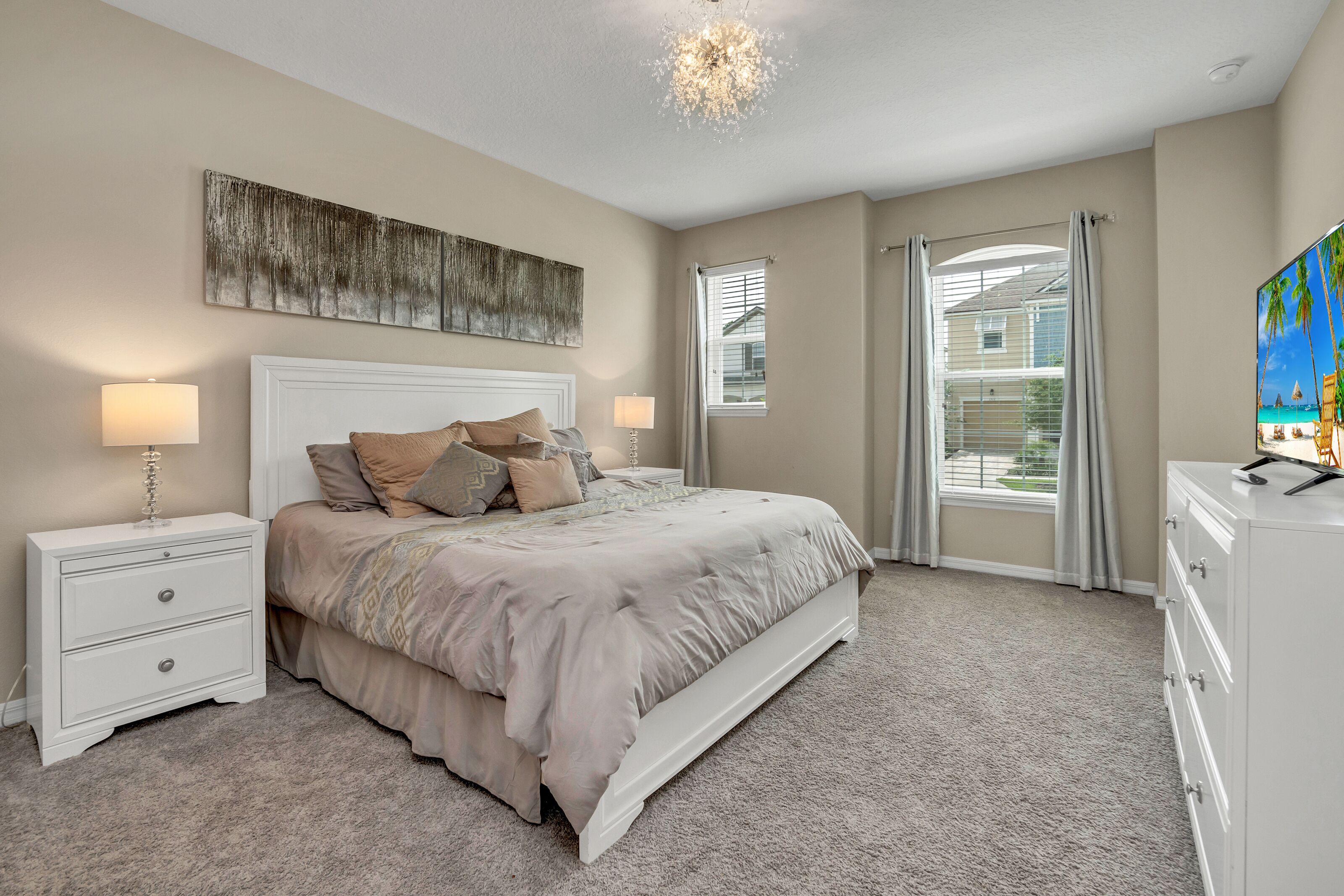 Une chambre à coucher avec un grand lit, deux tables de chevet, une commode, un lustre et une grande fenêtre avec des rideaux.