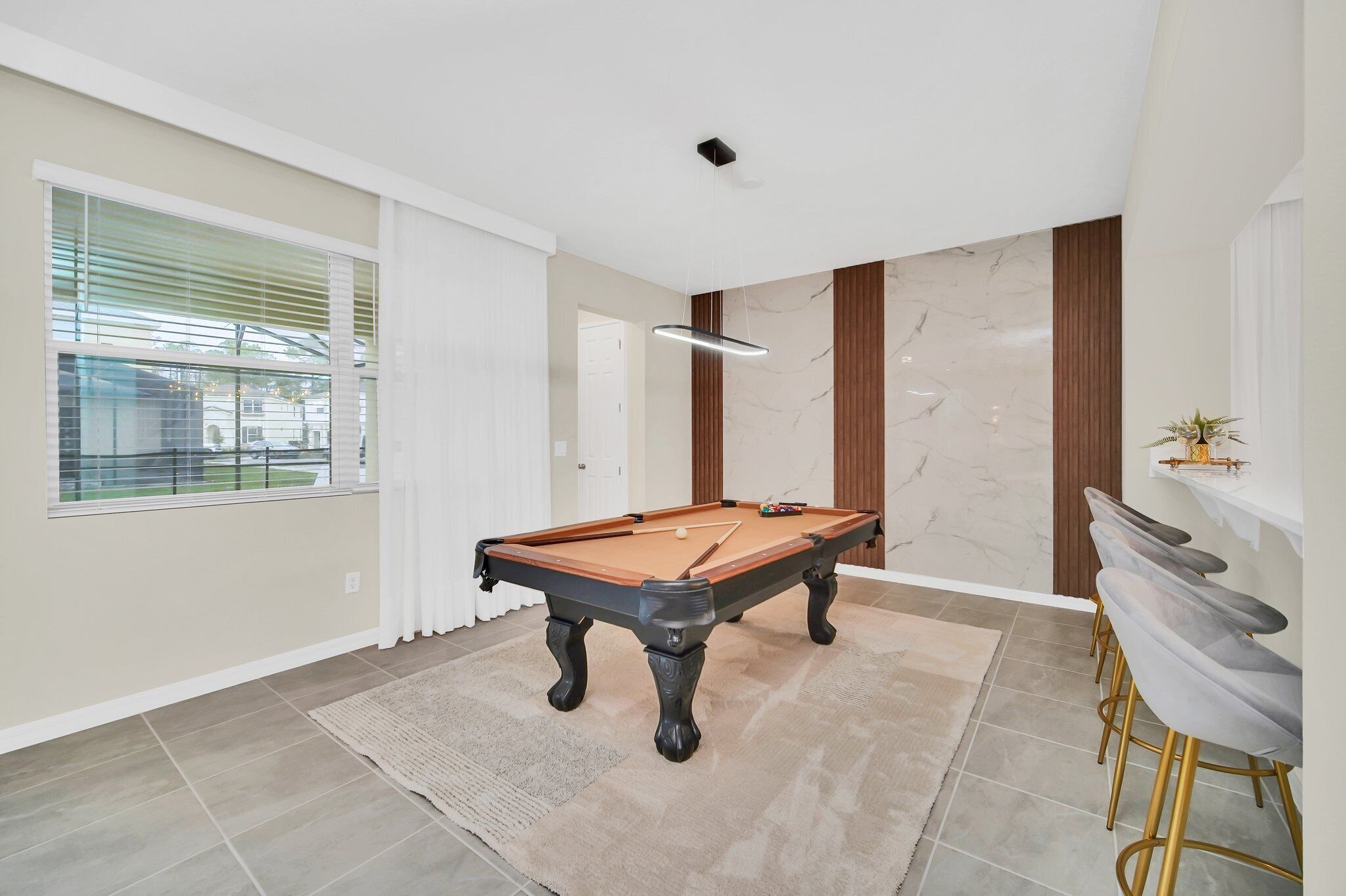 A modern living room with a pool table, a large window with blinds, a marble wall, and a bar area with stools.