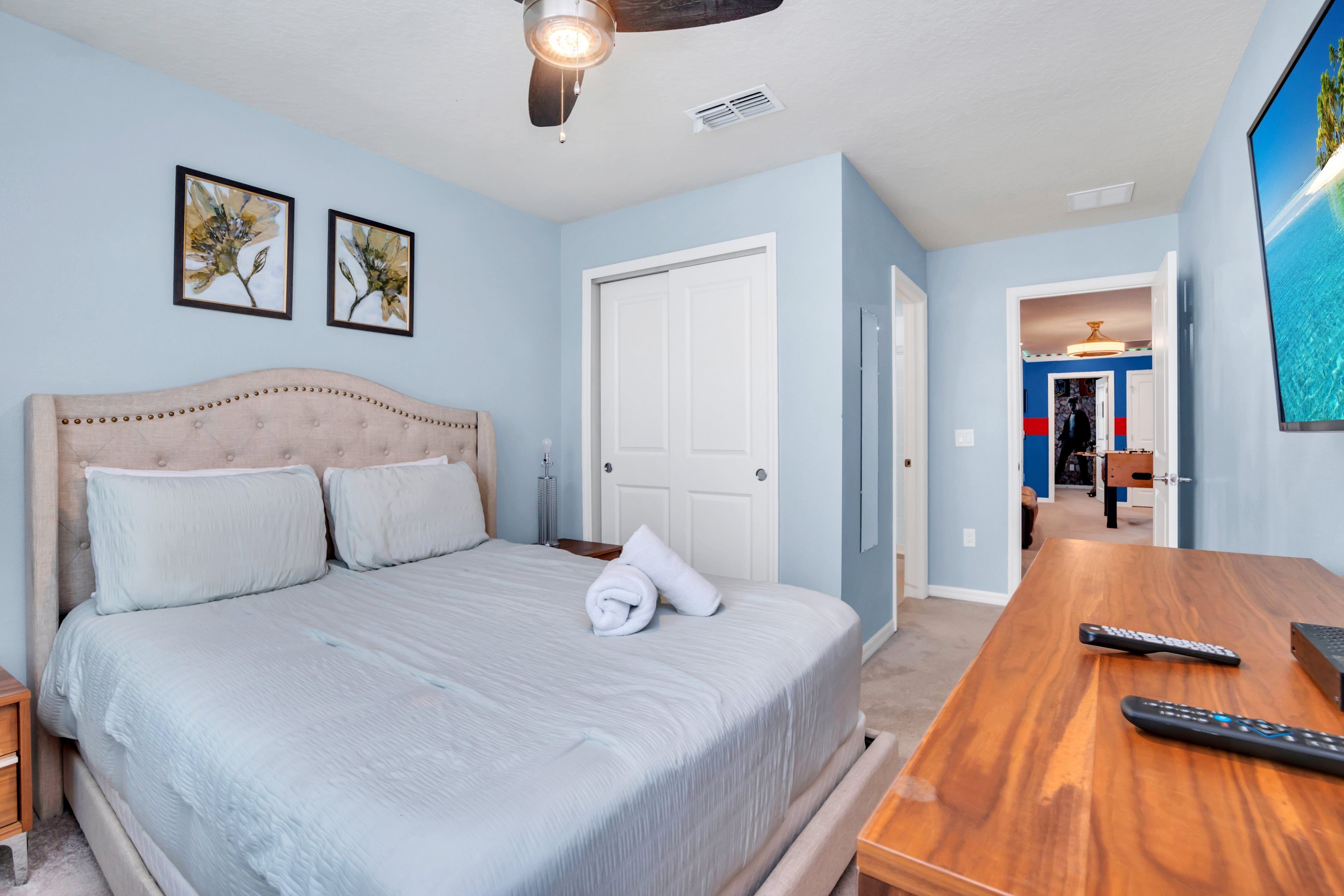 A bedroom with a bed, a wooden nightstand, a ceiling fan, and a TV on the wall.
