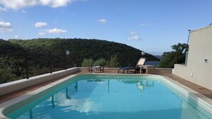 A rectangular swimming pool with a curved edge, surrounded by a wooden deck. There are two lounge chairs and a small table near the pool.