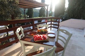 Une table à manger avec des chaises, un panier de fruits et des assiettes, le tout sur un balcon offrant une vue sur les arbres et une pergola.