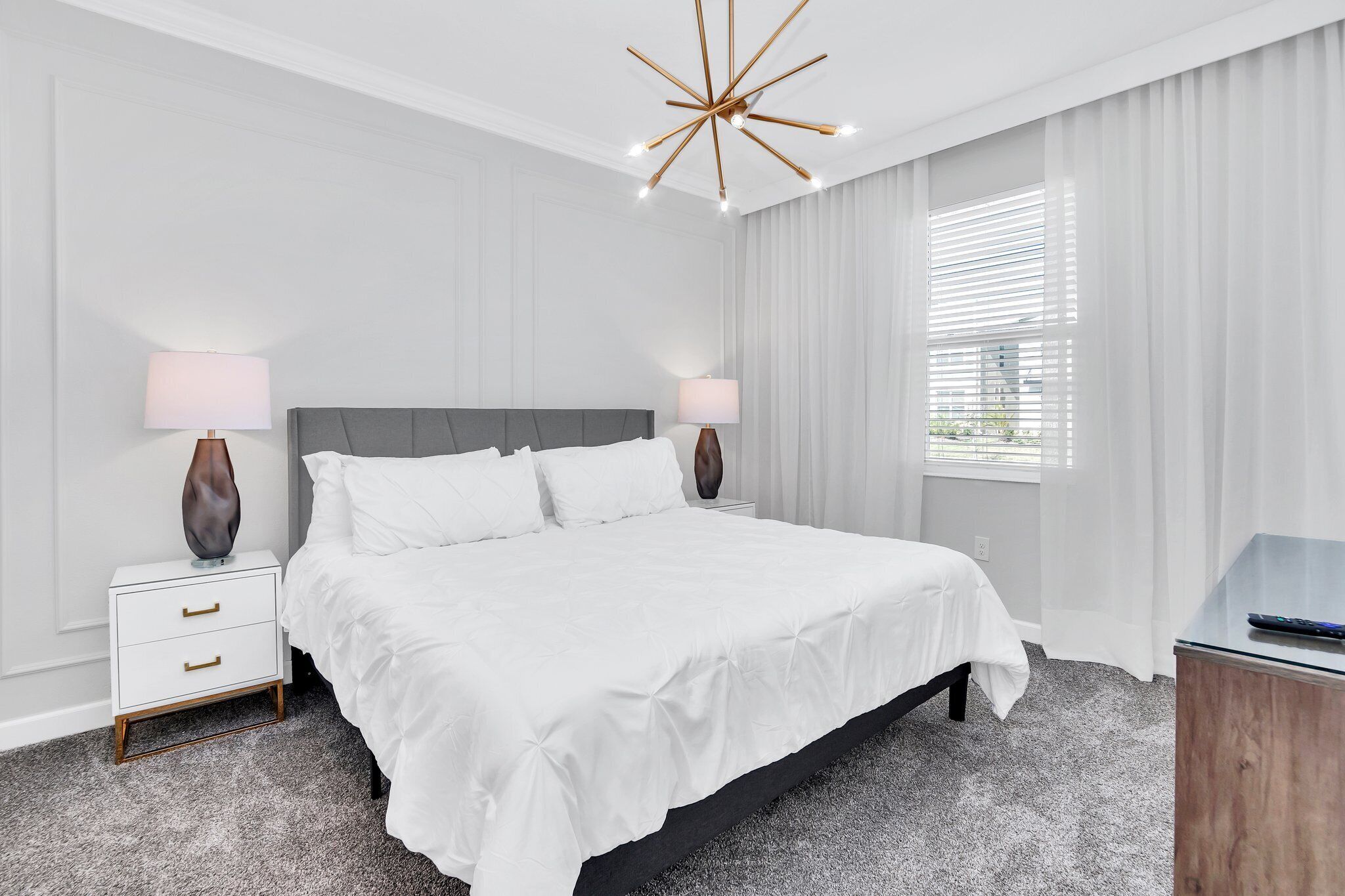 Une chambre d’hôtel moderne avec un grand lit, deux tables de chevet avec des lampes, un lustre contemporain et une vue dégagée à travers des rideaux légers.