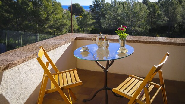 A glass-top table with two wooden chairs, a teapot, a cup, and a vase with flowers on a balcony with a view of trees and the sea.