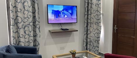 Una sala de estar con un televisor de pantalla plana montado en la pared, una mesa de centro de madera con cubierta de vidrio, dos sillones y cortinas estampadas.