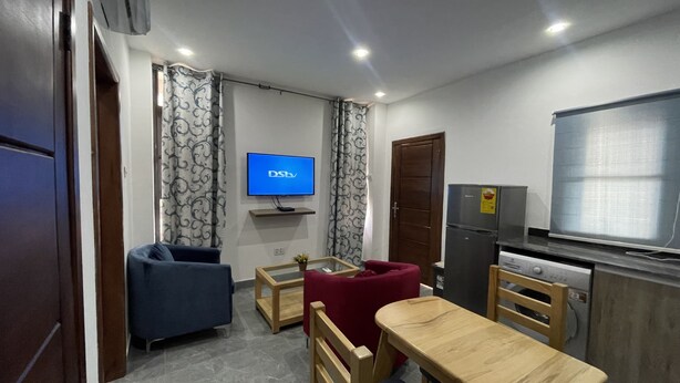 A compact living space with a television mounted on the wall, a refrigerator, a dining table with chairs, and a sofa. - AMERICAN MALL (Accra)