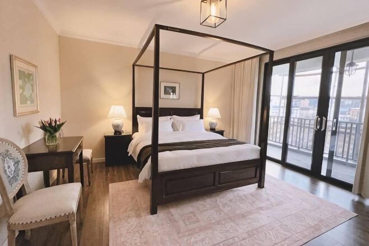 A four-poster bed with a nightstand, a chair, a desk, and a view of the outdoors through a sliding glass door.