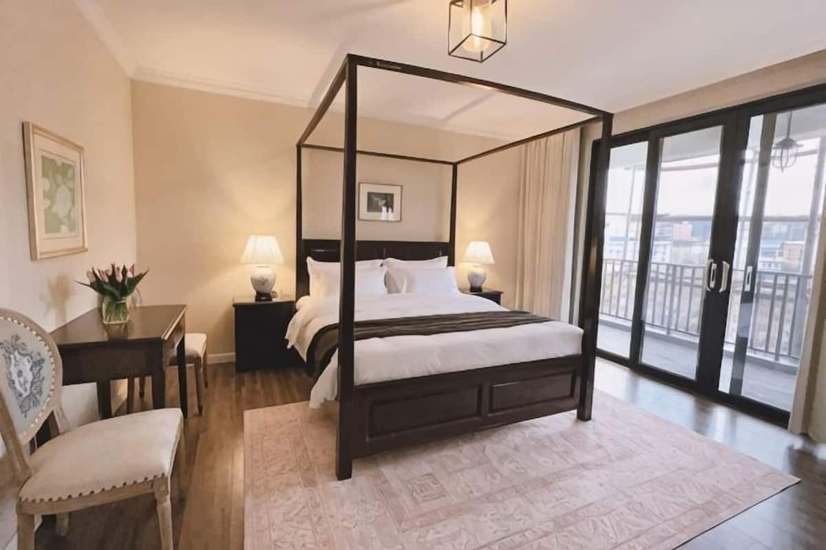 A four-poster bed with a nightstand, a chair, a desk, and a view of the outdoors through a sliding glass door.