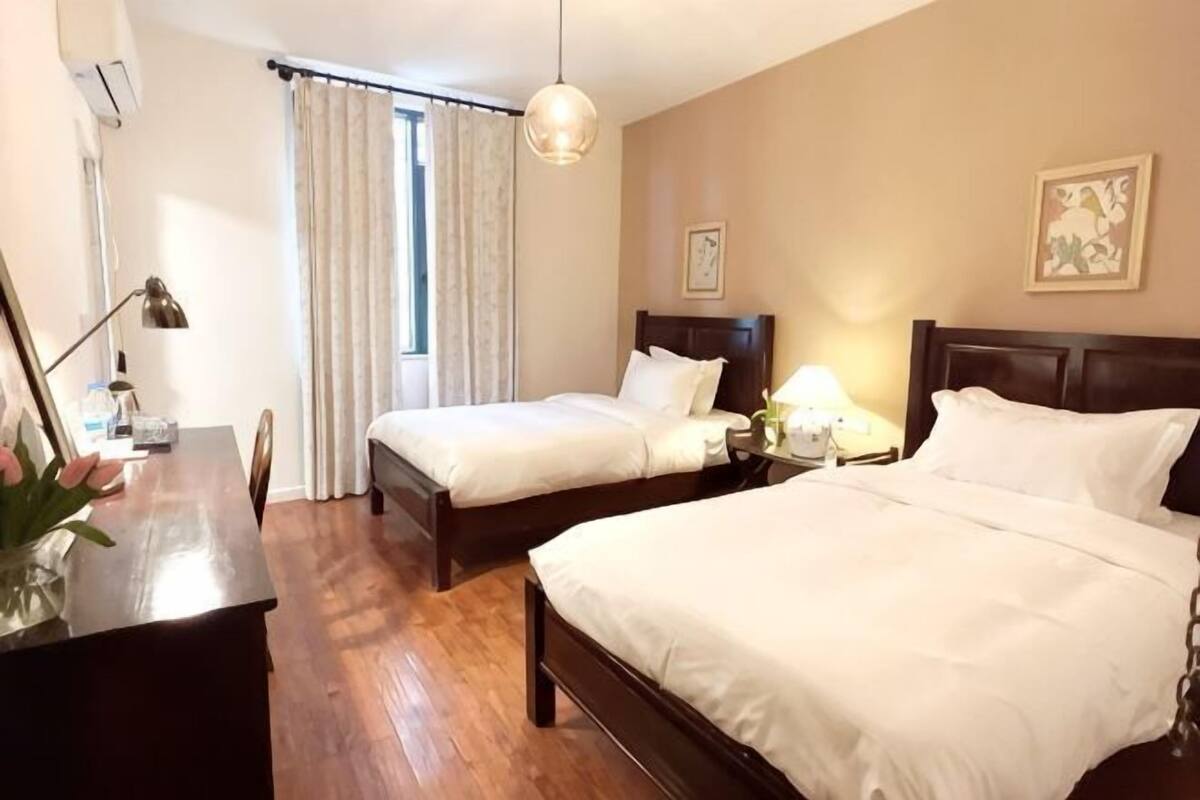 A hotel room with two beds, a desk, a chair, a lamp, a window with curtains, and a wall-mounted picture.