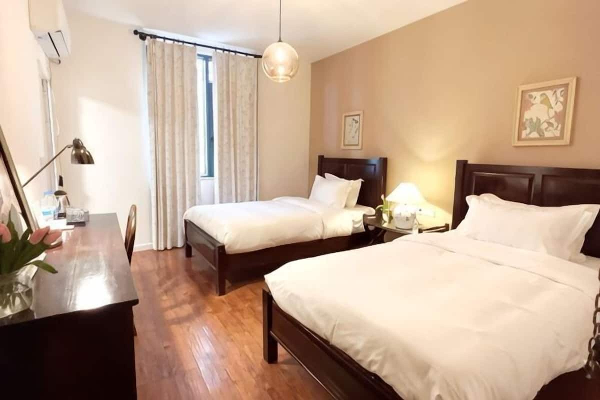 A hotel room with two beds, a desk, a chair, a lamp, a window with curtains, and a wall-mounted picture.