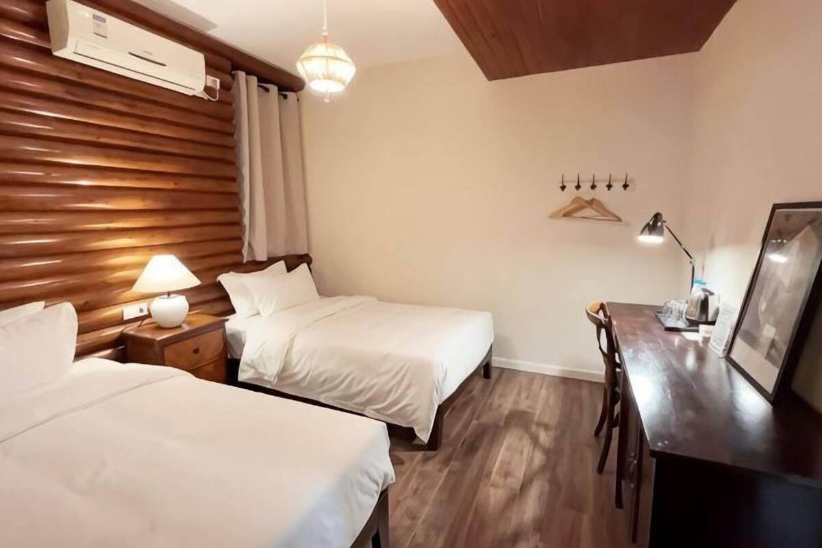A hotel room with two beds, a desk, a hanging light fixture, and a wall-mounted air conditioner.