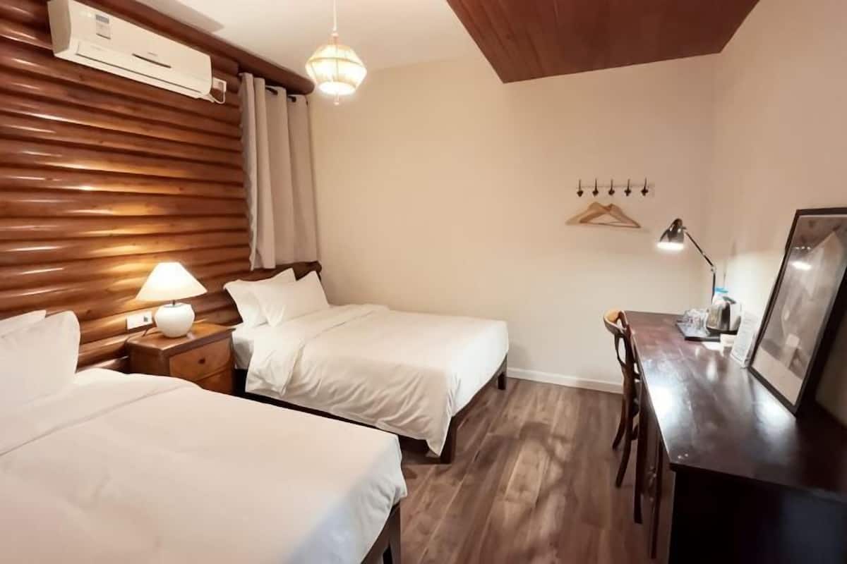 A hotel room with two beds, a desk, a hanging light fixture, and a wall-mounted air conditioner.