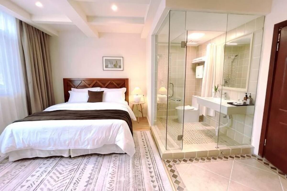 A hotel room with a bed, a glass-enclosed shower, a bathroom with a sink and mirror, and a door.