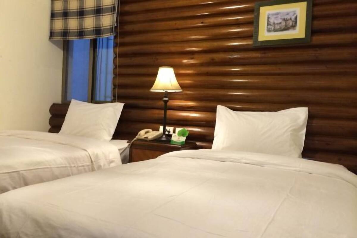 A hotel room with two beds, a wooden headboard, a nightstand with a lamp, a telephone, and a framed picture on the wall.