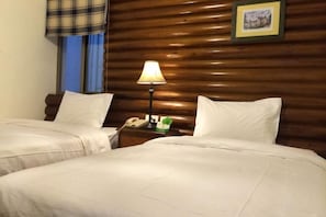 Una habitación de hotel con dos camas, cabecero de madera, mesita de noche con lámpara, teléfono y un cuadro enmarcado en la pared.