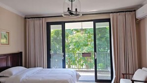 Habitación de hotel con cama, una silla, balcón con cortinas y vista a follaje.