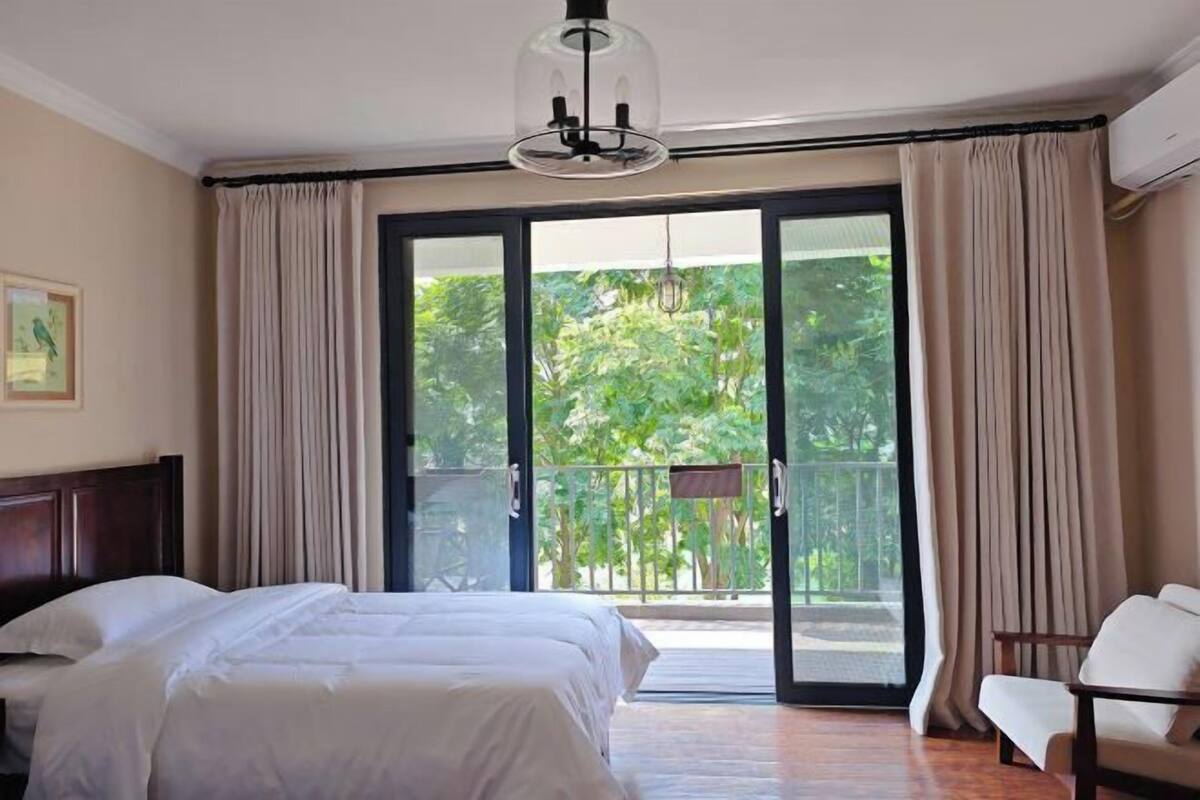 A hotel room with a bed, a chair, a balcony with curtains, and a view of greenery.