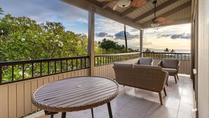 A covered balcony with wicker furniture, a round table, and a view of greenery and the sea. - Panoramic Ocean View Condo on Golf Course close to Beaches (Kihei)