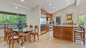 A modern kitchen with a central island, wooden chairs, and a glass dining table. - Panoramic Ocean View Condo on Golf Course close to Beaches (Kihei)