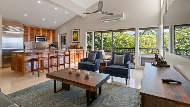 A modern kitchen with a central island, bar stools, a dining area with a wooden table, and a large window with a view of greenery. - Panoramic Ocean View Condo on Golf Course close to Beaches (Kihei)