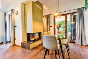 Relax in this stylish living room with a fireplace, glass dining table, and comfortable seating, offering a serene view of the lush garden.