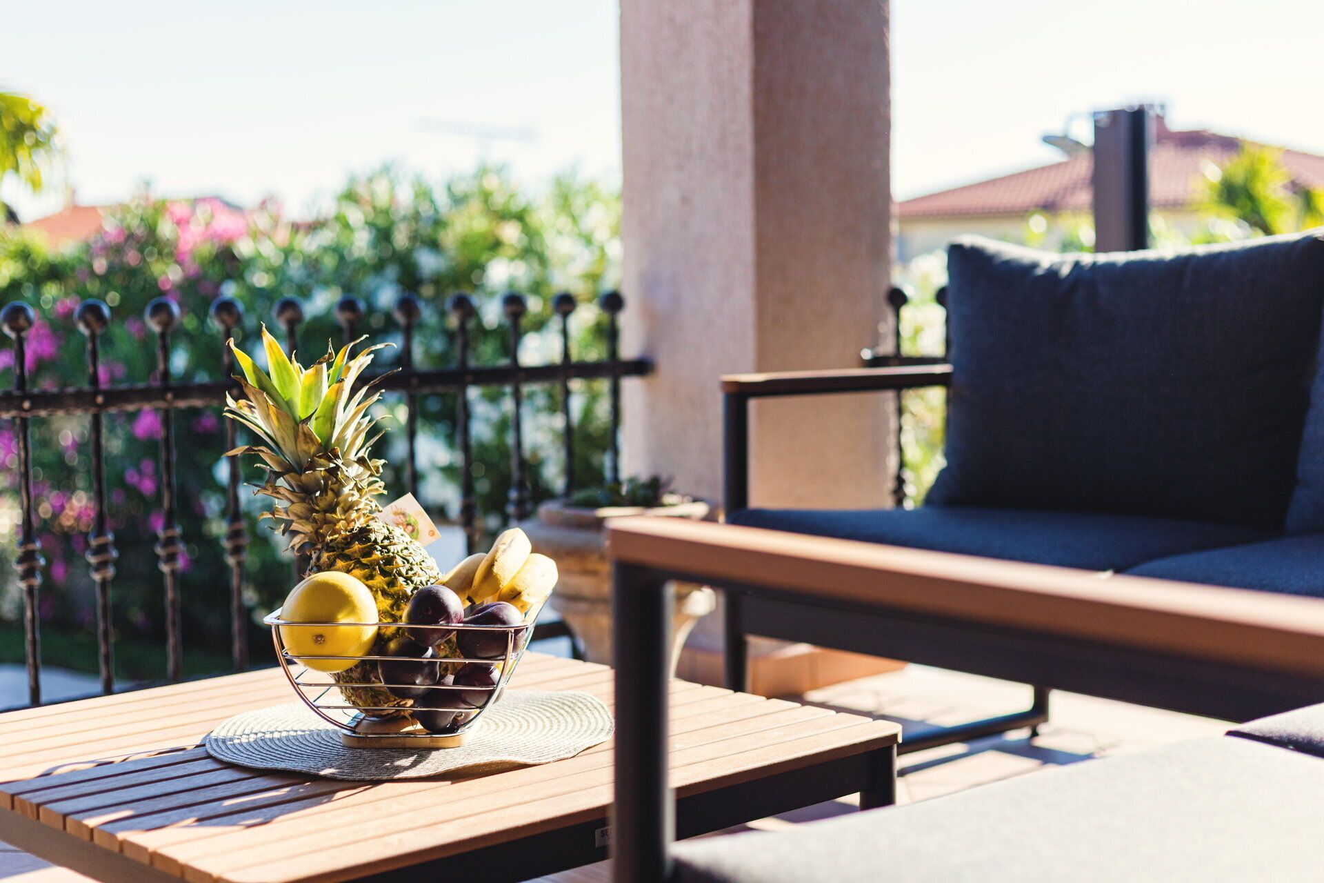 A wooden table with a fruit bowl containing pineapple, lemons, and plums, a blue cushioned chair, and a metal fence with greenery in the background.