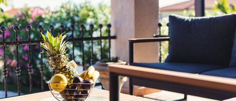A wooden table with a fruit bowl containing pineapple, lemons, and plums, a blue cushioned chair, and a metal fence with greenery in the background.