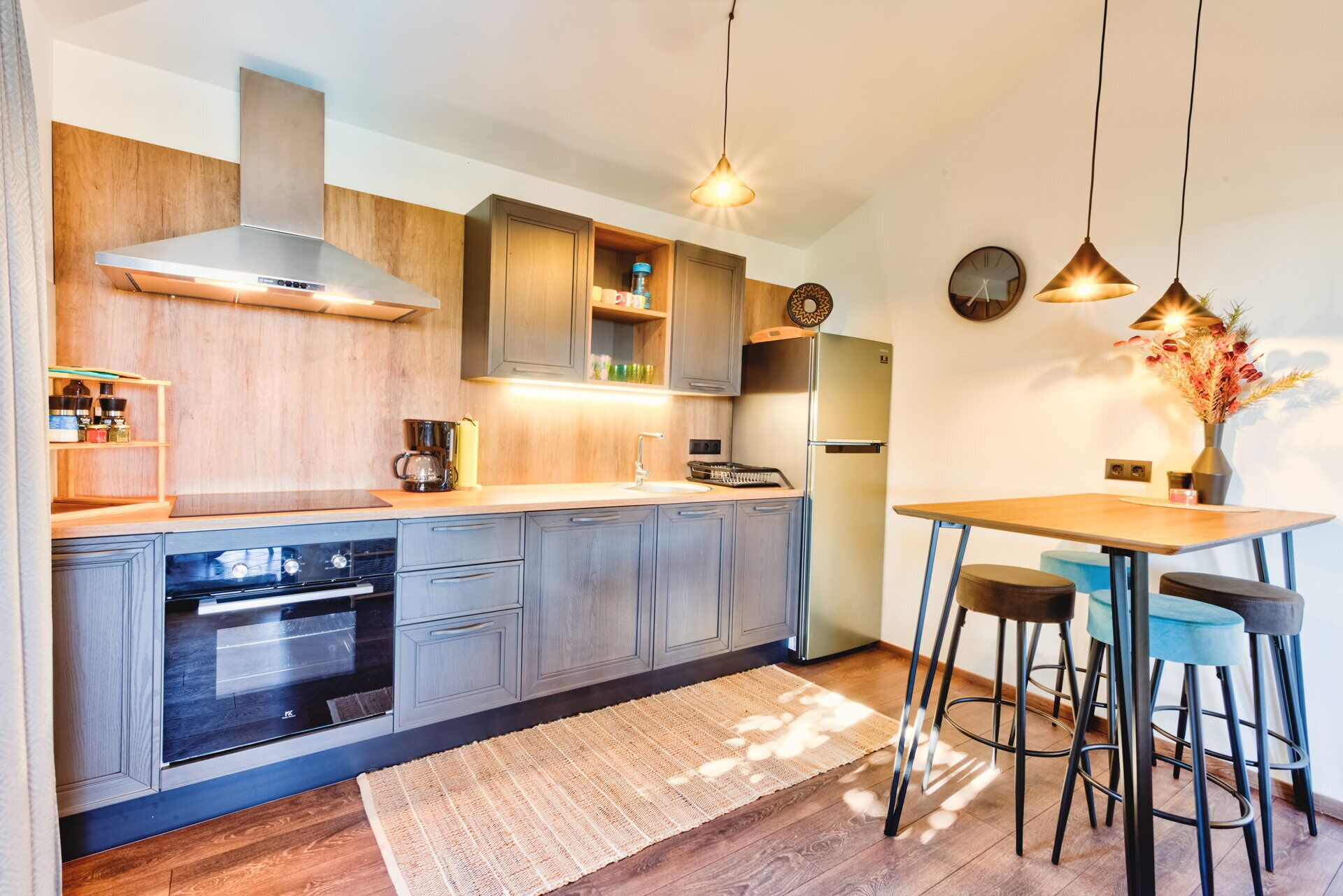 Een moderne keuken met houten kastjes, een roestvrijstalen afzuigkap, een ingebouwde oven, een koelkast, en een eethoek met krukken.