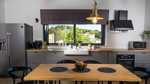 Een moderne keuken met een houten eettafel, zwarte stoelen, een roestvrijstalen koelkast, een koffiezetapparaat en uitzicht op groen buiten.