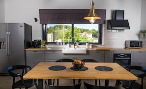 A modern kitchen with a wooden dining table, black chairs, a stainless steel refrigerator, a coffee maker, and a view of greenery outside.