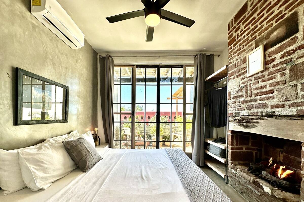 Ein Schlafzimmer mit einem großen Bett, einem Deckenventilator, einer Klimaanlage, einem Kamin aus Ziegeln und Blick auf ein Gebäude durch ein großes Fenster.