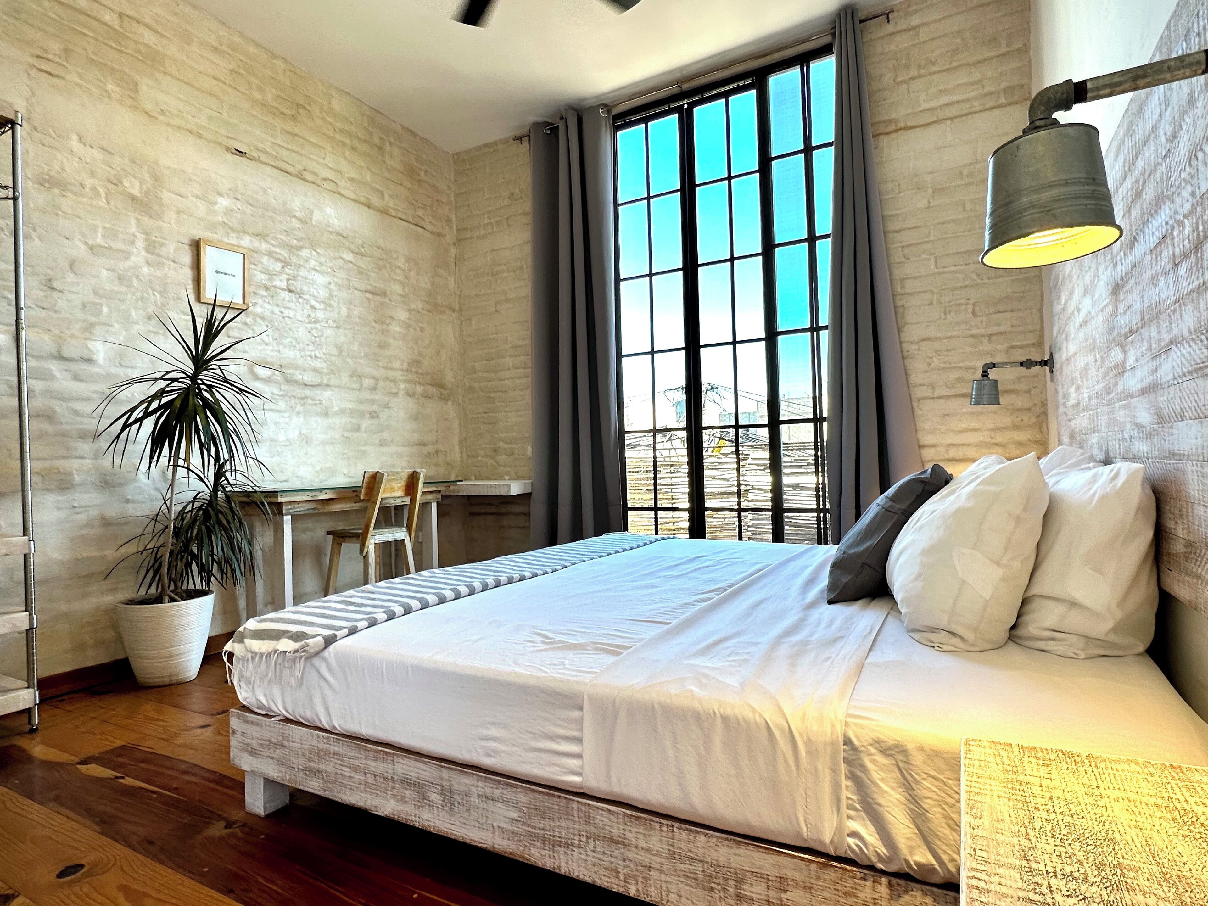 A bedroom with a large bed, a desk, a chair, a potted plant, and a window with curtains.